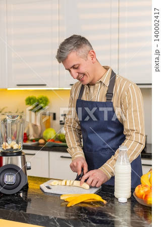 Middle-aged man slicing banana for blender in kitchen preparing smoothie for diet nutrition routine 140002417