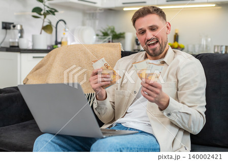 Young man counts cash euros compares with laptop earnings sheet fills sums smiles proud at goal met Young man counts cash euros compares with laptop earnings sheet fills sums smiles proud at goal met 140002421