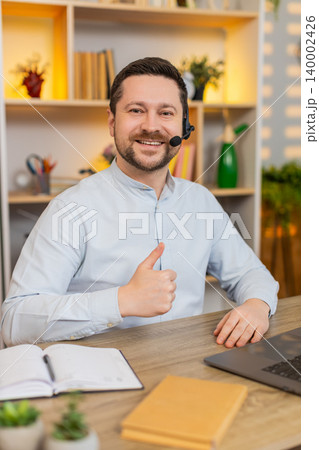 Young man on home table working in headset with laptop and smiling during virtual customer support Young man on home table working in headset with laptop and smiling during virtual customer support 140002426