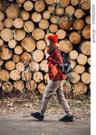 Woman walks past stacked timber logs in woodland area. Woman forest inspector, female industrial professional, environmental auditor at work. Woman walks past stacked timber logs in woodland area. Woman forest inspector, female industrial professional, environmental auditor at work. 140002669