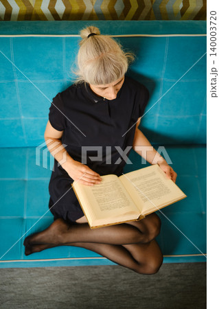 Person sits on couch and reads book in casual indoor setting during day Person sits on couch and reads book in casual indoor setting during day 140003720