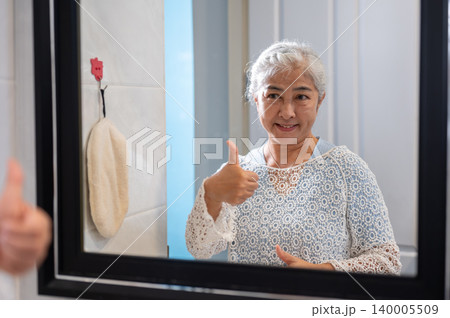 Happy elderly old asian woman grandma giving thumb up to herself as looking at reflection in mirror. Happy elderly old asian woman grandma giving thumb up to herself as looking at reflection in mirror. 140005509