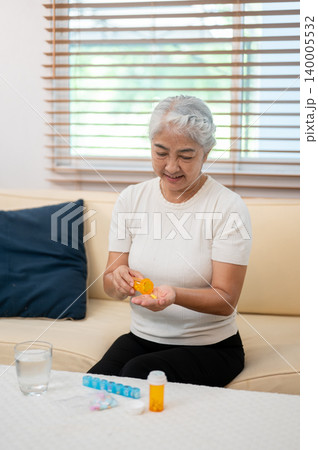 Elderly old asian woman grandma taking medicine or supplement while sitting on sofa in a living room Elderly old asian woman grandma taking medicine or supplement while sitting on sofa in a living room 140005532