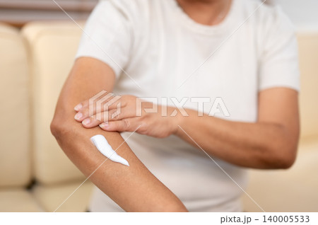 Close up of elderly old asian woman grandma applying skin lotion cream on arm while sitting on sofa. Close up of elderly old asian woman grandma applying skin lotion cream on arm while sitting on sofa. 140005533