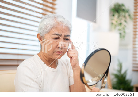 Elderly old asian woman grandma holding a round mirror looking at eyes wrinkle while sitting on sofa 140005551
