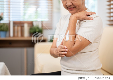 Close up of elderly old asian woman grandma applying skin lotion cream on arm as she sitting on sofa 140005563