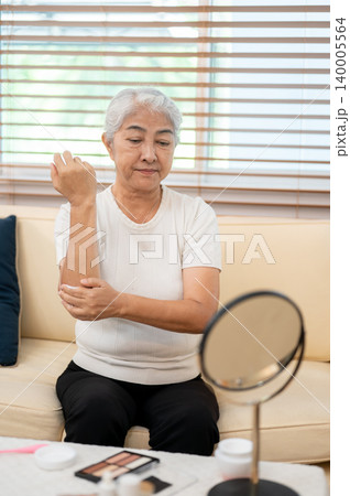 Elderly old asian woman grandma applying skin lotion cream on arm in front of mirror sitting on sofa Elderly old asian woman grandma applying skin lotion cream on arm in front of mirror sitting on sofa 140005564