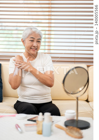 Elderly old asian woman grandma applying skin lotion cream looking in mirror as she sitting on sofa. Elderly old asian woman grandma applying skin lotion cream looking in mirror as she sitting on sofa. 140005566
