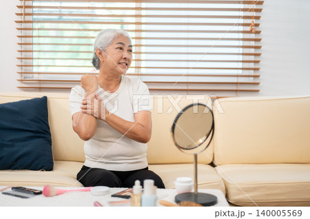 Happy elderly old asian woman grandma applying skin lotion cream in front of mirror sitting on sofa. Happy elderly old asian woman grandma applying skin lotion cream in front of mirror sitting on sofa. 140005569
