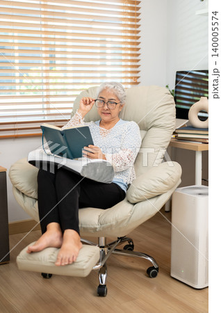 Full body of glasses elderly old asian woman reading a book on sofa armchair in living room at home. Full body of glasses elderly old asian woman reading a book on sofa armchair in living room at home. 140005574