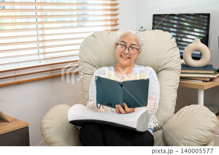 Glasses retired elderly old asian woman reading a book on comfy sofa armchair in living room at home Glasses retired elderly old asian woman reading a book on comfy sofa armchair in living room at home 140005577