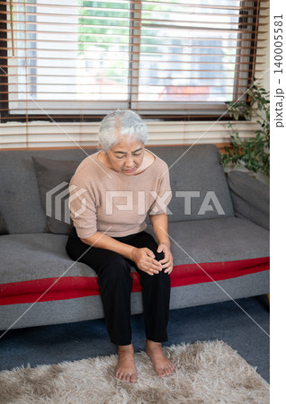 Elderly old asian woman grandma holding and looking at knee getting pain ache while sitting on sofa. Elderly old asian woman grandma holding and looking at knee getting pain ache while sitting on sofa. 140005581