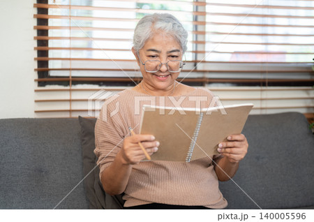 Smiling glasses old asian woman grandma reading notebook as sitting on sofa in living room at home. Smiling glasses old asian woman grandma reading notebook as sitting on sofa in living room at home. 140005596