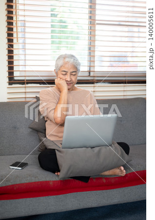 Sleepy old asian woman grandma closing eyes over laptop sitting folding legs on sofa in living room Sleepy old asian woman grandma closing eyes over laptop sitting folding legs on sofa in living room 140005611