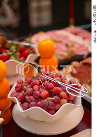 Bowl of red grapes with serving tongs, surrounded by stacked oranges, strawberries, cheese, and charcuterie on a banquet table. Bowl of red grapes with serving tongs, surrounded by stacked oranges, strawberries, cheese, and charcuterie on a banquet table. 140005980
