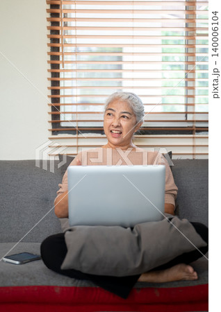 Happy old asian woman grandma looking over laptop while sitting folding legs on sofa in living room. 140006104