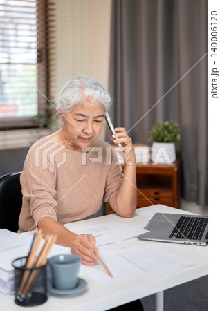 Senior old asian woman holding pencil talking on phone aside laptop at working table in home office. 140006120