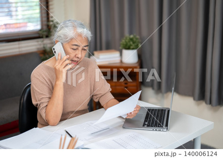 Elderly senior old asian woman holding paper document talking on phone call at table in home office. 140006124