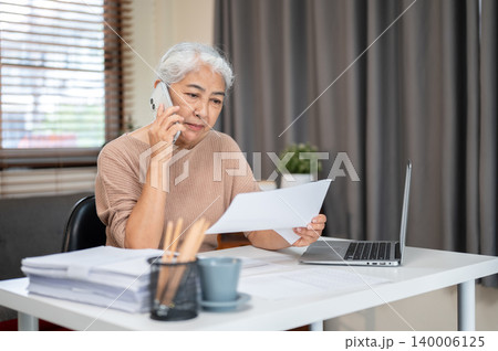 Elderly senior old asian woman holding reading document as talking on phone at table in home office 140006125