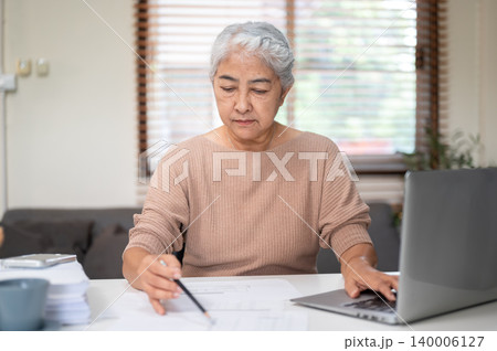 Senior old asian woman holding pencil writing on paper aside laptop at working table at home office. 140006127