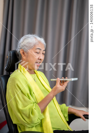 Elderly old asian woman holding phone talking on call with speaker on chair in living room at home. 140006138