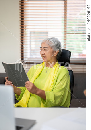 Smiling elderly old asian woman reading a book while sitting on chair in living room at home office. 140006160