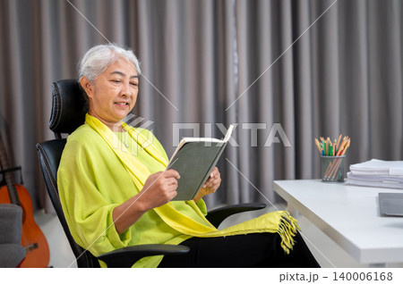 Happy elderly old asian woman reading a book while sitting leaning on chair in a living room at home Happy elderly old asian woman reading a book while sitting leaning on chair in a living room at home 140006168