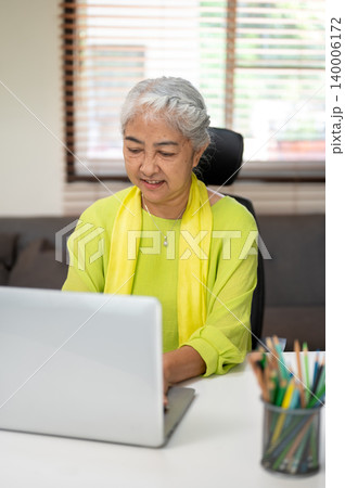 Smart elderly senior old asian woman typing on laptop while sitting at working table in home office. Smart elderly senior old asian woman typing on laptop while sitting at working table in home office. 140006172