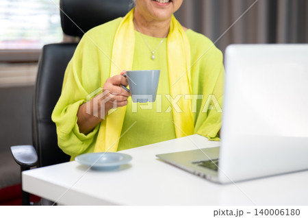 Close up of smiling elderly old asian woman holding coffee looking at laptop on table in home office Close up of smiling elderly old asian woman holding coffee looking at laptop on table in home office 140006180