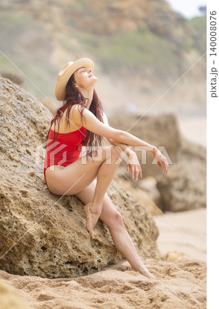 Woman sits on a rock at the beach during a sunny day and enjoys the warm weather while wearing a swimsuit and hat Woman sits on a rock at the beach during a sunny day and enjoys the warm weather while wearing a swimsuit and hat 140008076