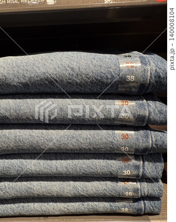 Stack of blue denim jeans on display shelf in retail store with size labels visible 140008104