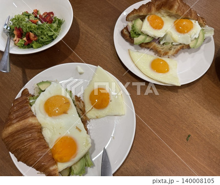Breakfast croissant sandwich with fried eggs and avocado on white plates with fresh salad added. 140008105
