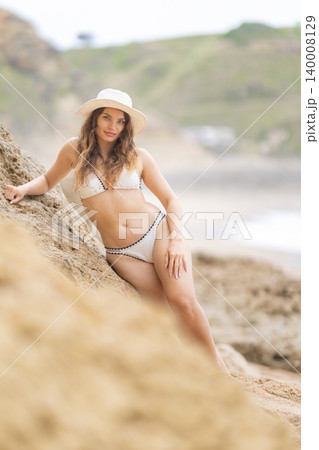 Woman in white bikini poses on sandy beach near rocky cliffs during sunny day 140008129
