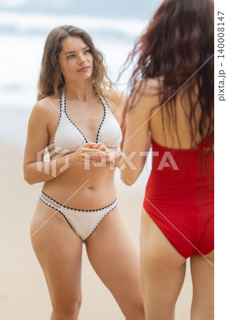 Two women chat and enjoy the beach on a sunny day while wearing swimwear and standing on the sand near the ocean Two women chat and enjoy the beach on a sunny day while wearing swimwear and standing on the sand near the ocean 140008147