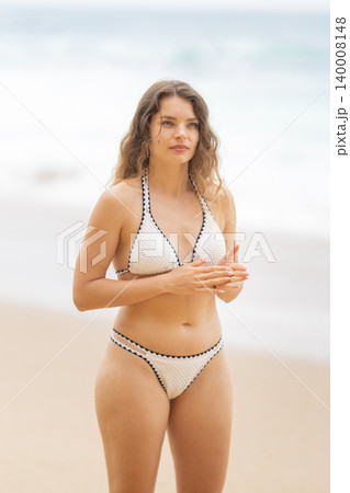 Woman stands at the beach wearing a bikini by the ocean during a sunny day in daylight hours 140008148