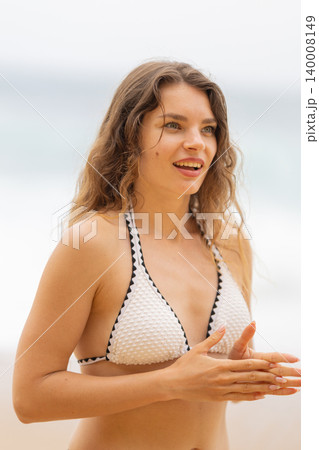 Woman smiles while standing on the beach during a sunny day with waves in the background and warm weather 140008149