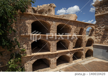 A row of empty arched brick crypts and stone burial niches is visible in a historic Spanish cemetery. The sky above is blue with some clouds. 140010420