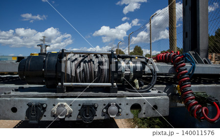An electric towing winch is mounted on an empty transport trailer. The trailer is placed outdoors under a clear blue sky with white clouds in the background. 140011576