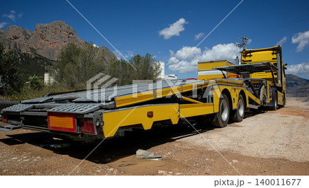 A yellow car carrier truck is parked on a dirt road. The truck is located near rocky mountains and surrounded by trees. The sky is clear with few clouds. A yellow car carrier truck is parked on a dirt road. The truck is located near rocky mountains and surrounded by trees. The sky is clear with few clouds. 140011677