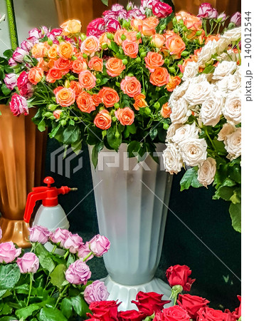 Colorful bouquet of roses in various shades arranged in a tall white vase with a spray bottle visible among additional flowers in a floral shop setting Colorful bouquet of roses in various shades arranged in a tall white vase with a spray bottle visible among additional flowers in a floral shop setting 140012544