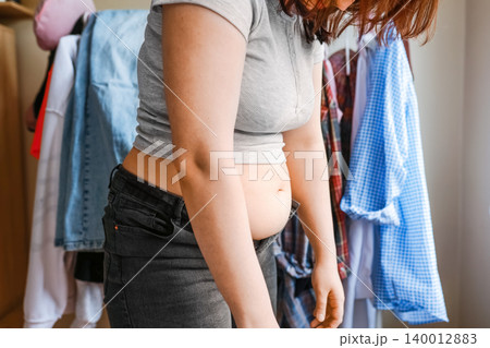 An overweight woman stands by her wardrobe with an exposed belly and unbuttoned tight jeans, frustrated by weight gain and clothing annoyance in a plus-size lifestyle. An overweight woman stands by her wardrobe with an exposed belly and unbuttoned tight jeans, frustrated by weight gain and clothing annoyance in a plus-size lifestyle. 140012883