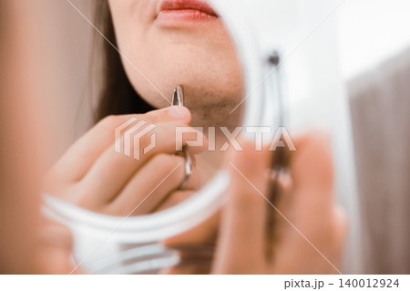 Reflection in a mirror of a woman using tweezers for facial hair removal on her chin, managing unwanted hair and hormonal changes in her home beauty routine. 140012924