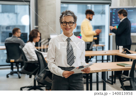 Confident businesswoman in formal attire holding documents, smiling at camera in modern office environment with colleagues engaged in discussions and teamwork Confident businesswoman in formal attire holding documents, smiling at camera in modern office environment with colleagues engaged in discussions and teamwork 140013958