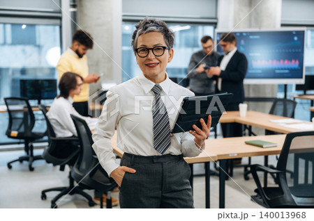 Confident businesswoman holding tablet in modern office, surrounded by colleagues engaged in discussions, showcasing teamwork and collaboration in a professional environment Confident businesswoman holding tablet in modern office, surrounded by colleagues engaged in discussions, showcasing teamwork and collaboration in a professional environment 140013968