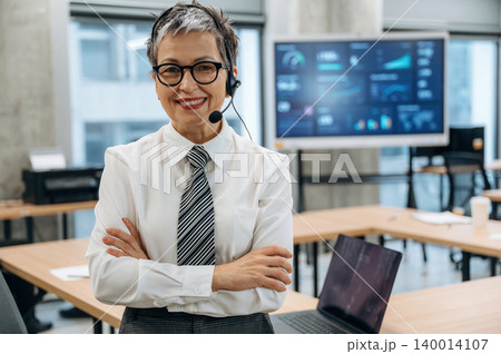 Confident businesswoman in a modern office setting, wearing a headset and smiling, showcasing professionalism and teamwork in a collaborative environment 140014107