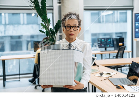 Businesswoman carrying a box of office supplies and a plant in a modern workspace, showcasing organization and productivity in a professional environment Businesswoman carrying a box of office supplies and a plant in a modern workspace, showcasing organization and productivity in a professional environment 140014122