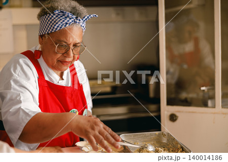 Senior Black woman in her sixties preparing food in a professional kitchen wearing red apron. Senior Black woman in her sixties preparing food in a professional kitchen wearing red apron. 140014186