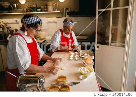 Active Senior Hispanic and Black women rolling dough and baking in a professional community kitchen Active Senior Hispanic and Black women rolling dough and baking in a professional community kitchen 140014190