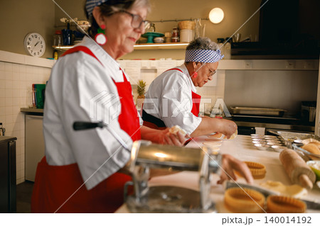 Active senior Caucasian and Black female chefs making pastries in a professional kitchen setting. 140014192