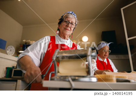 Senior Caucasian woman and African American friend making fresh pasta in a kitchen workshop 140014195